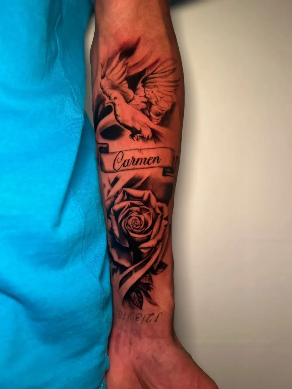 Memorial forearm tattoo: a dove over a banner reading Carmen, a rose below